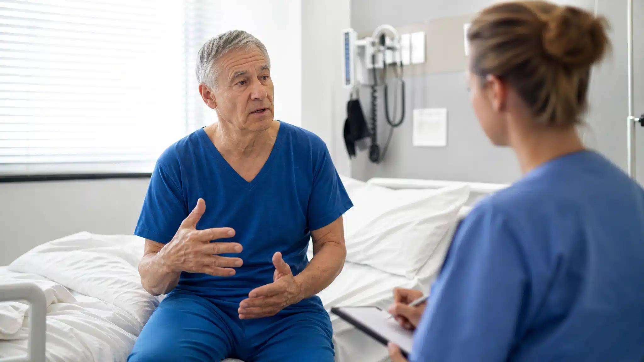 Older man in medical scrubs speaking with a healthcare professional while sitting on a hospital bed